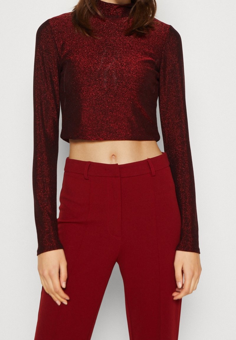 Roter glitzernder, kürzerer Langarm-Top mit hohem Kragen, kombiniert mit maßgeschneiderten roten Hosen. Beide Kleidungsstücke haben eine glatte Textur und eine elegante Passform.