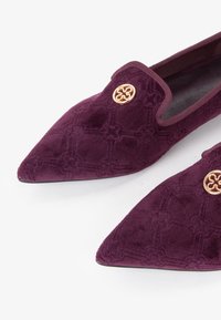 Un paio di ballerine a punta in velluto viola scuro con sottili motivi geometrici cuciti e accenti decorativi circolari dorati sulla parte superiore.