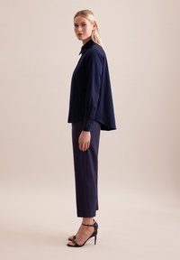 Granatowa koszula zapinana na guziki z długimi rękawami, gładki materiał i zaokrąglony dół, w zestawie z dopasowanymi granatowymi spodniami oraz czarnymi sandałami na wysokim obcasie.
