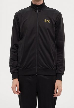 Mężczyzna w czarnej kurtce zapinanej na zamek z długimi rękawami i żółtym logo EA7 Emporio Armani na klatce piersiowej.