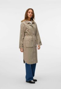 Beige vattentät jacka med knappstängning, elastisk midja, två framfickor och sidoflikar, i kombination med vida blå jeans och svarta skor.