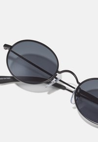 Round sunglasses with dark lenses, thin black metal frame, clear nose pads, and straight temples. Minimalist design with no visible logos.