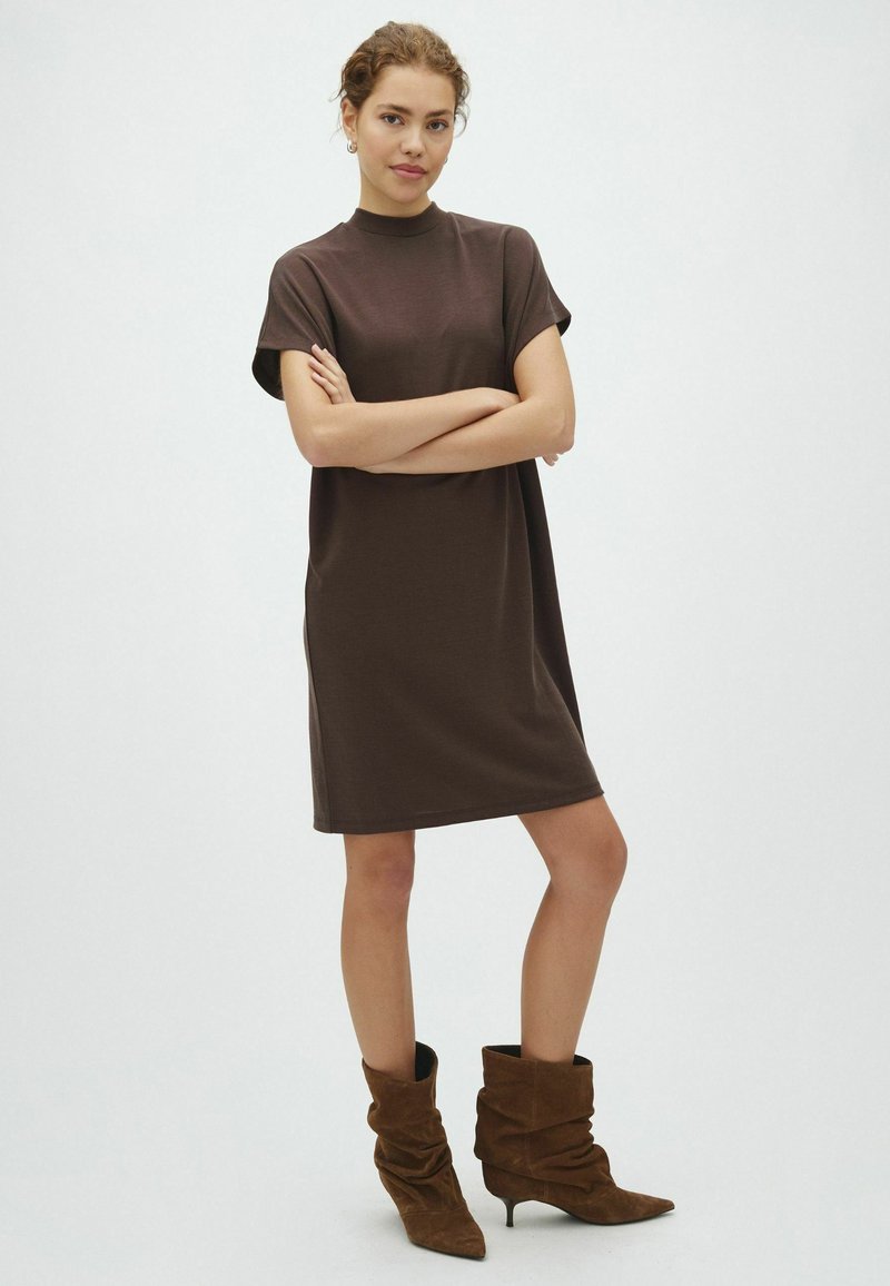 Femme debout, les bras croisés, portant une robe marron à manches courtes et des bottines marron décontractées, sur un fond clair uni.