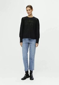 Pull en maille noire avec un design perforé et des manches longues, associé à un jean bleu clair et des bottines noires brillantes.