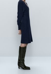 Robe tricotée bleu marine avec un col en V et des manches longues, accompagnée de bottes en cuir noir jusqu'aux genoux avec des pointes et un talon fin.