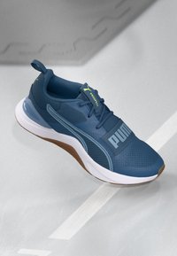 Blå idrottssko med en texturerad meshovandel, med en framträdande grå "PUMA"-logotyp och en vit sula med en brun gummidetalj.