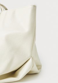 Kremowa torba typu tote o miękkiej, teksturowanej powierzchni, charakteryzująca się wyraźnym kształtem i subtelnymi fałdami u podstawy.