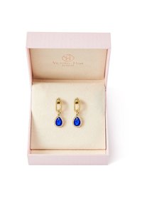 Pendientes de oro con piedras en forma de lágrima azul, rodeados de pequeñas gemas transparentes, presentados en una caja de color rosa pálido con un interior suave y claro.
