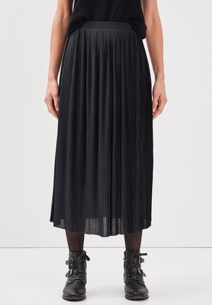 Vrouw draagt een zwarte geplooide midi-rok met zwarte panty's en zwarte veterlaarzen met gespsdetails, staand tegen een effen achtergrond.