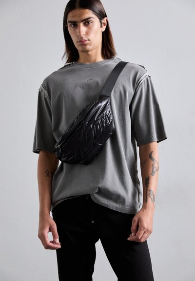 Junger Mann mit langen Haaren, der ein graues, übergroßes T-Shirt, schwarze Hosen, eine schwarze Umhängetasche trägt und sichtbare Tattoos an beiden Unterarmen hat.