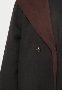 Abrigo de lana negro y marrón con un gran cuello, presenta un botón negro y una textura suave. Diseño entallado con un frente abierto.