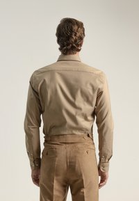 Camisa beige de manga larga con diseño de espalda recta, que cuenta con cuello y puños con botones, combinada con pantalones de tiro alto en color marrón claro.