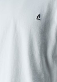 Valkoinen puuvillapaita pyöreällä kaula-aukolla ja vasemmassa rinnassa pienellä mustalla liekkilogolla brodeerattuna. Sileä pinta, ei näkyviä kuvioita.