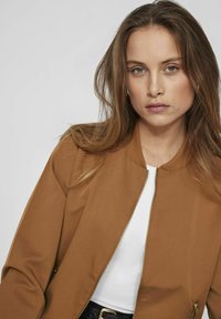 Hnedý bombr jacket s hladkou textúrou, zipsom vpredu a ribbed golierom; nosený na bielom topu, doplnený čiernym textúrovaným opaskom.