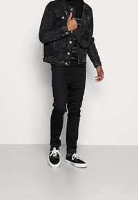 Giubbotto di denim nero con una finitura leggermente sbiadita, indossato sopra un maglione nero, abbinato a jeans neri slim e sneakers nere con suole bianche.