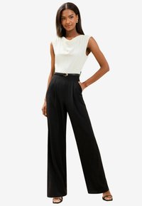 White cowl neck top with sleeveless design, paired with high-waisted black wide-leg trousers. Black belt with gold buckle accent.