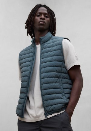 Ung mand med dreadlocks iført en blå quiltet vest over en hvid T-shirt, stående med den ene hånd i lommen mod en ensfarvet baggrund.