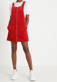 Rotes Corduroy-Latzkleid mit einem Reißverschluss vorn, Trägern und seitlichen Taschen, getragen über einem weißen Rippenshirt und kombiniert mit weißen Sneakern.