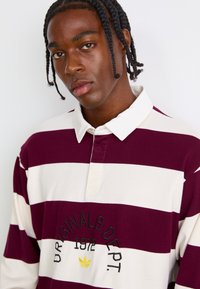 Burgundi és fehér csíkos, hosszú ujjú pólóing, hegyes gallérral és hímzett logóval. Puha tapintás, figyelemre méltó varrási részletekkel.