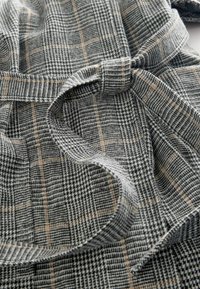 Ruuduline kangas halli, musta ja beeži toonides. Pehme tekstuur ja seotud vöö detail. Kujunduses kasutatakse koertehammas- ja ruudumustreid visuaalse huvi loomiseks.