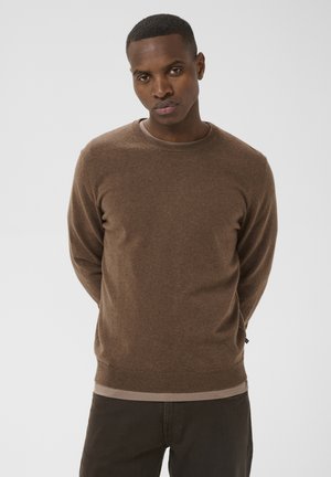 Homme portant un pull marron à manches longues, debout avec les mains derrière le dos, devant un fond clair uni.