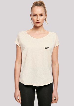 Light beige short-sleeve t-shirt with a round neckline. Features a black "SLAY" text detail on the chest. Soft, textured fabric.