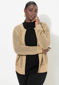 Cardigan lavorato a maglia beige con un design in rete leggero, chiusura a bottoni, polsini a coste e una vestibilità morbida su un top a collo alto nero.