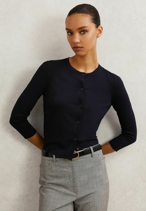 Jeune femme aux cheveux lissés en arrière porte un cardigan noir boutonné avec des manches trois-quarts et un pantalon gris à carreaux avec une ceinture noire.