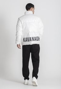 Hvid puffjakke med høj krave og ryglogo "KAVANAGH." Kombineret med sorte bukser og hvide sneakers. Glat, skinnende tekstur.