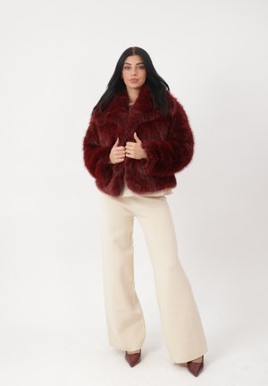 Frau mit langen dunklen Haaren, die eine weinrote Kunstpelzjacke, cremefarbene Hosen mit weitem Bein und weinrote spitze Pumps trägt, stehend vor weißem Hintergrund.