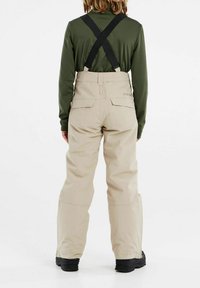 Beige Schneehosen mit verstellbaren schwarzen Trägern, weitem Schnitt und zwei Gesäßtaschen, getragen über einem eng anliegenden grünen Langarmshirt.