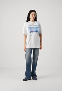 Mulher de pé, vestindo jeans e uma t-shirt oversized branca com a estampa "Palm Springs", segurando uma bolsa de ombro azul clara contra um fundo simples.