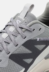 Gros plan d'une chaussure de sport grise avec des lacets à motifs, des accents en suède, des panneaux en mesh et une languette de tirage.