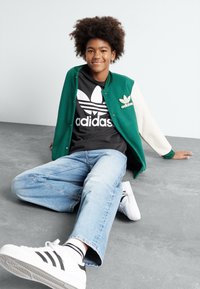 Zaļš un krēmkrāsas varianto žakets virs melnas Adidas T-kreklam, apvienots ar gaiši ziliem džinsiem un baltiem apaviem ar melnām svītrām.