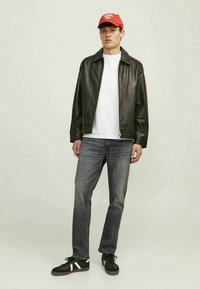 Casaco de couro preto, t-shirt branca, jeans cinzentos e ténis pretos com riscas brancas. Boné de basebol vermelho com detalhe em texto. Fundo neutro.