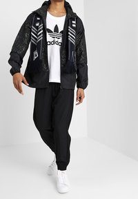 Svart jacka med mönstrade ärmar, vit t-shirt med svart logotyp, lösa svarta byxor, vita sneakers och en marinblå scarf med ränder och fransar.