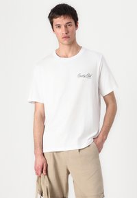 Witte katoenen T-shirt met korte mouwen, voorzien van een zwart 'Country Club' logo op de linkerkant van de borst, gecombineerd met beige shorts.