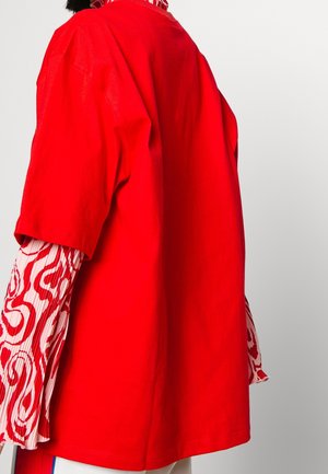 Persoon draagt een effen felrode oversized T-shirt over een rood-wit patroon met lange mouwen, gezien van achteren en opzij.