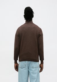 Brūns adīts džemperis ar augstu apkakli, teksturētu virsmu un ribojošu malu, kombinēts ar gaiši ziliem džinsa biksēm.