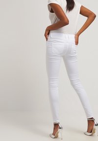 Vaqueros skinny blancos de denim, con una textura suave, bolsillos traseros y un ajuste ceñido. Combinados con una blusa de color claro y tacones metálicos.