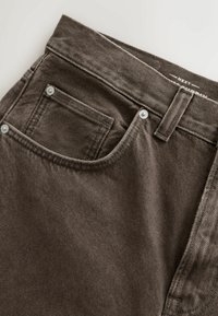 Közelkép barna farmernadrág zsebével, látható szegecsekkel, övbújtatóval és részben olvasható derékpántcímkével, melyen a "NEXT" felirat szerepel.