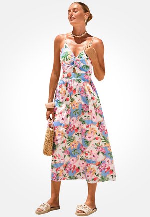 Žena u midi cvjetnoj haljini bez rukava sa tanke naramenice, drži pletenu torbu, stoji sa zatvorenim očima i opuštenim izrazom lica.