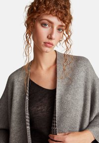 Šedý pletený cardigan s měkkou texturou, který má kontrastní vzorovaný lem podél otevřeného předního dílu a uvolněný střih.