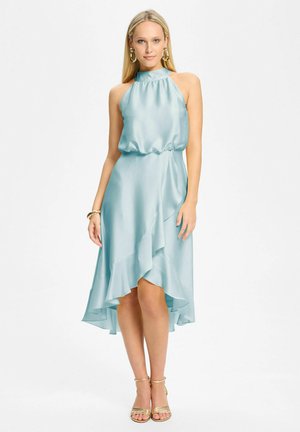 Femme blonde portant une robe sans manches en satin bleu clair à ourlet haut-bas avec volant, des sandales dorées et de grandes boucles d'oreilles créoles dorées, debout.