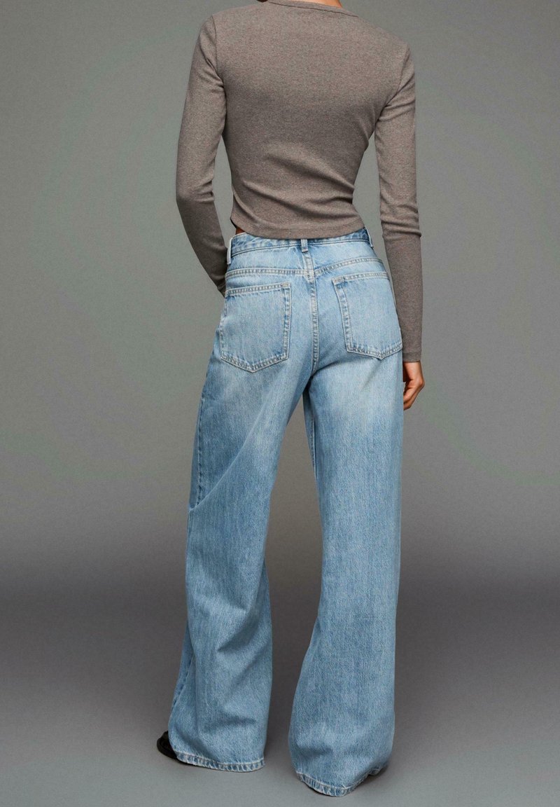 Personne debout, de dos, portant un haut gris ajusté à manches longues et un jean large bleu clair sur un fond uni.