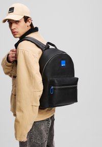 Black backpack with textured leather, rounded shape, front zip pocket with blue pull tab, worn by a person in a tan jacket and cap.