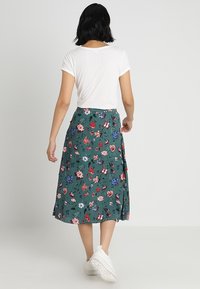 Blommig grön midiklänning med rosa, röda och blå blommor, med en flödande design, mjukt tyg och dragkedja bak. Matchas med vita sneakers.