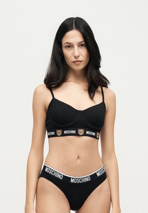 Un mannequin aux longs cheveux foncés porte un bralette noir Moschino et une culotte assortie sur un fond clair uni.