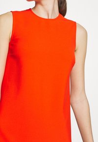 Bezrukávové oranžové šaty s kulatým výstřihem, hladkým materiálem a těsným střihem, s čistými liniemi a bez viditelných vzorů nebo dekorací.