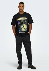 Camiseta de algodón negra con un estampado gráfico de SpongeBob y el texto "Rock Bottom". Combinada con pantalones cargo negros y zapatos negros.